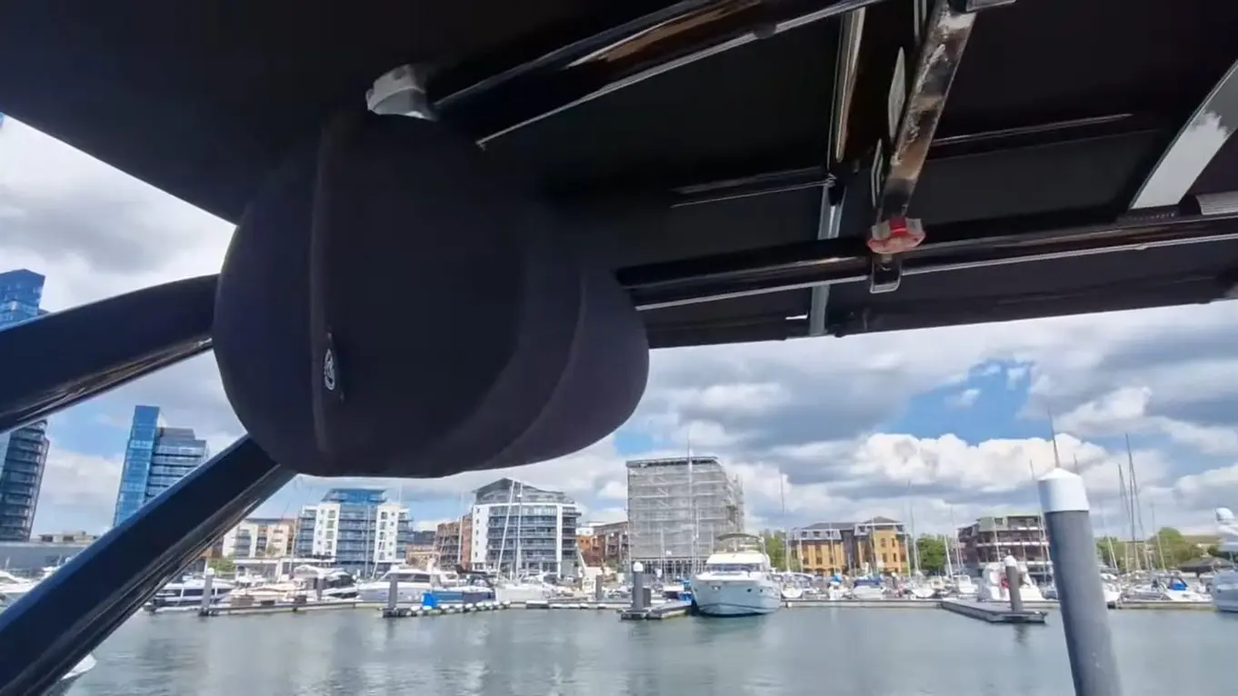 View of a marina with various boats, featuring a black speaker hanging from a yacht's roof against a blue sky.